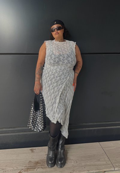 Vestido blanco de encaje con un diseño texturizado y en capas. Bolso tote negro con tachuelas, botas negras altas hasta el muslo. La modelo lleva gafas de sol y una gorra.