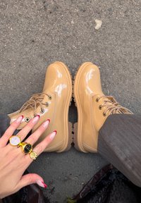 Botas bege em couro verniz com solas em borracha texturizada, biqueira arredondada e ilhós em metal. Mão com unhas bem cuidadas e anéis em primeiro plano.