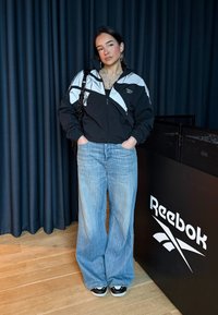 Femme portant une veste Reebok noire et blanche, un jean bleu large et des baskets noires, debout les mains dans les poches près d'un panneau portant la marque Reebok.