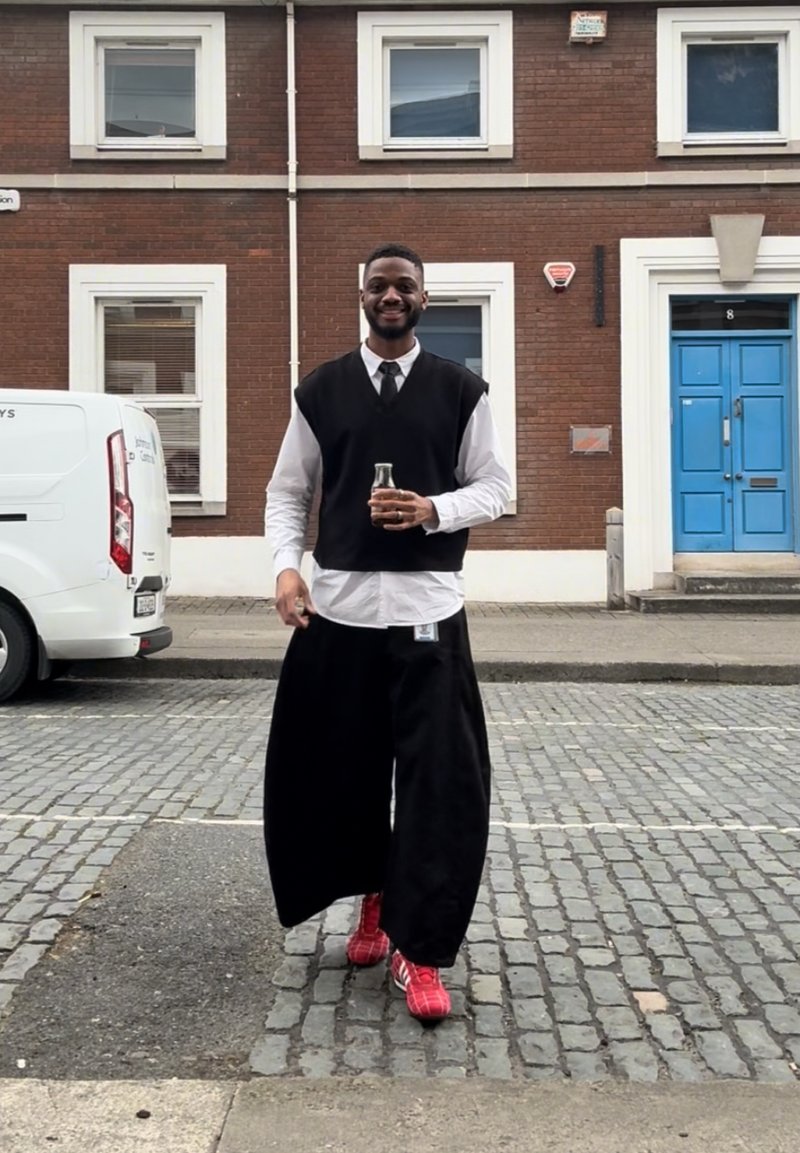 Pantalon noir oversize, pull sans manches noir porté par-dessus une chemise blanche, baskets rouges, et une bouteille en verre. Rue pavée en arrière-plan.