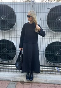 Čierny skrátený sveter s dlhými rukávmi, skombinovaný s čiernou maxi sukňou. Modelka nosí tkanú čiernu tašku a má na sebe čierne členkové čižmy.