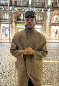 Homme portant un manteau marron à carreaux, une casquette noire et des lunettes, debout les mains jointes devant un bâtiment en pierre éclairé avec un panneau "PALACE".