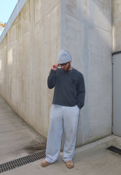 Hombre con gorro gris, sudadera oscura, pantalones deportivos gris claro y gafas, de pie contra una pared de cemento cerca de una puerta al aire libre.