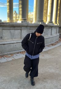 Μαύρο bomber μπουφάν με ασημένιους φερμουάρ, φορεμένο πάνω από ένα μπλε καρό πουκάμισο και χαλαρούς μαύρους παντελόνες. Συνδυασμένο με καπέλο beanie και μαύρα μποτάκια.