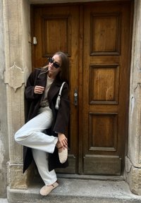 Hnědý oversized kabát, bílá košile a kalhoty. Texturované béžové boty. Stojí u dřevěných dveří s dekorativními panely a kamenou zdí.