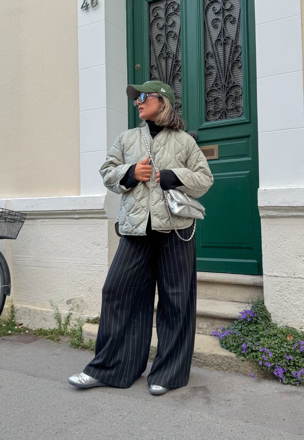 Quilted silver jacket, black turtleneck, wide black striped trousers, silver trainers, metallic handbag, and an olive cap in front of a green door.