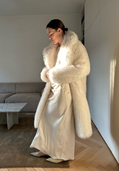 Abrigo de piel sintética blanca con mangas largas sobre un vestido satinado crema, combinado con unos tacones puntiagudos en verde claro, posando frente a un fondo minimalista.