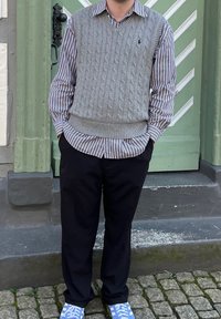 Hallitus halli lõngakootud kampsun triibulise särgi peal, koos mustade pükste ja siniste tossudega. Taustal roheline uks.