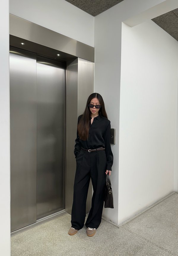 Vrouw in zwarte outfit en zonnebril staat bij gesloten roestvrijstalen liftdeuren en houdt een zwarte handtas vast in een modern gebouw.