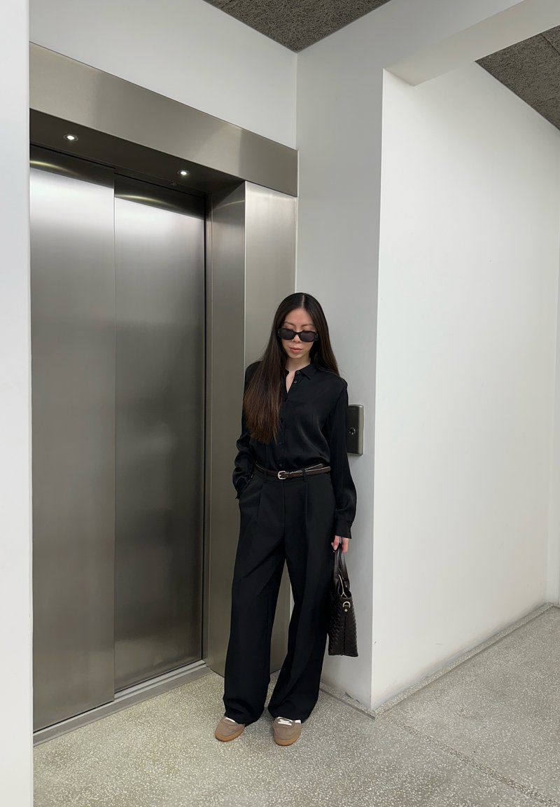 Frau in schwarzem Outfit und Sonnenbrille steht vor geschlossenen Aufzügen aus Edelstahl und hält eine schwarze Handtasche in einem modernen Gebäude.