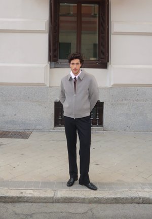 Siv zip-up jopič nad belo srajco z burgundskim kragnim, črnimi hlačami in čevlji. Oseba stoji na kamnitem hodniku blizu okna.