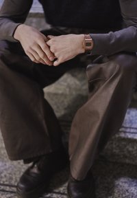 Personne portant une chemise à manches longues foncée et un pantalon en cuir marron, assise sur des marches en pierre, les mains jointes, avec une montre numérique rose doré au poignet.