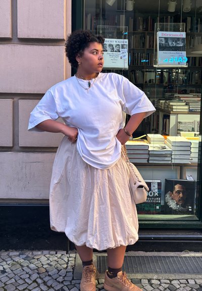 Camiseta blanca de gran tamaño, falda vaporosa color crema, zapatillas beige y un bolso ligero con correa de cadena. Libros al fondo.