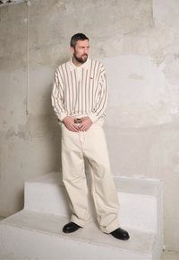 Camisa polo riscada em creme com mangas longas, combinada com calças largas bege e sapatos pretos, em pé em degraus brancos contra uma parede texturizada.