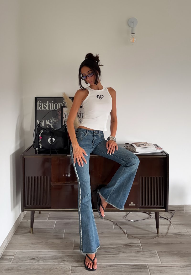 Femme portant un top blanc sans manches et un jean évasé, posant avec une jambe levée dans une pièce avec un meuble en bois et des objets décoratifs derrière elle.