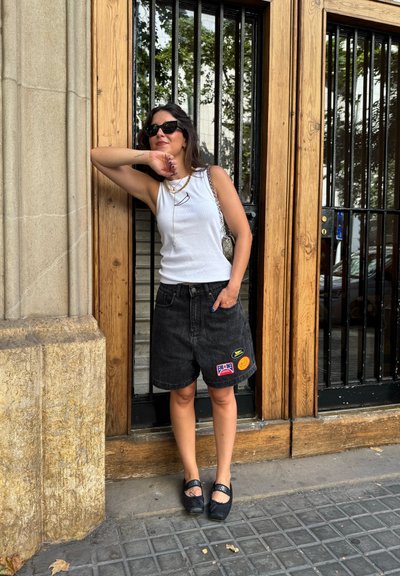 Shorts de mezclilla negra con parches, top blanco de tirantes, collares superpuestos y bailarinas negras con acentos decorativos, frente a puertas de madera.