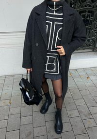 Cappotto di lana grigio con grandi bottoni, maglione a fantasia in bianco e nero, gonna grigia, collant neri trasparenti, stivaletti neri in pelle alla caviglia e una borsa.