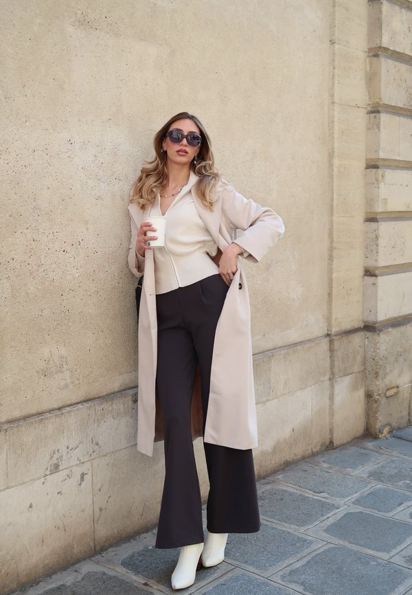Femme en trench-coat beige, haut blanc à fermeture éclair, pantalon noir à jambes larges et bottes blanches, debout contre un mur en pierre, tenant une tasse de café, portant des lunettes de soleil.