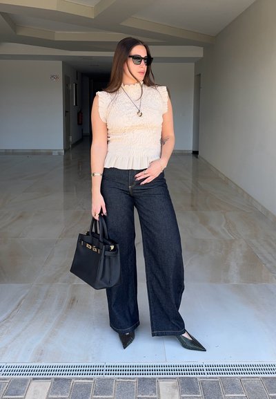 Mujer con gafas de sol, top blanco sin mangas con volantes, vaqueros oscuros de pierna ancha, zapatos puntiagudos con tachuelas, sosteniendo bolso negro en interiores.