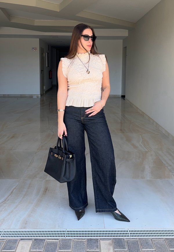 Žena nosící sluneční brýle, bílý bezrukávový top s volánky, široké tmavé džíny, špičaté boty s cvočky, držící černou kabelku v interiéru.
