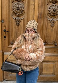 Veste en fausse fourrure de couleur marron clair avec des taches blanches, col montant et fermeture éclair. Bonnet beige à pois marron. Sac bandoulière noir.