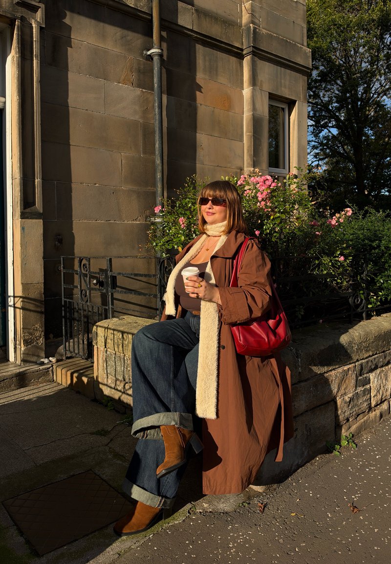 Une femme portant des lunettes de soleil est assise près d’un bâtiment en pierre et tient un café à emporter. Elle a un sac rouge en bandoulière, et des fleurs roses se trouvent derrière elle.