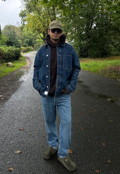 Chaqueta de denim sobre una sudadera marrón, jeans de color azul claro y zapatos slip-on verdes. Los accesorios incluyen una gorra de camuflaje y gafas de sol.