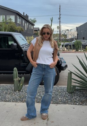 Mujer de cabello rubio con gafas de sol, camiseta blanca, chaleco marrón, vaqueros azules y zapatos marrones de pie en la acera cerca de un Jeep negro y cactus.