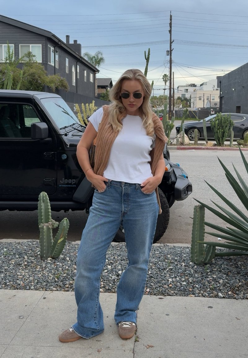 Mujer de cabello rubio con gafas de sol, camiseta blanca, chaleco marrón, vaqueros azules y zapatos marrones de pie en la acera cerca de un Jeep negro y cactus.