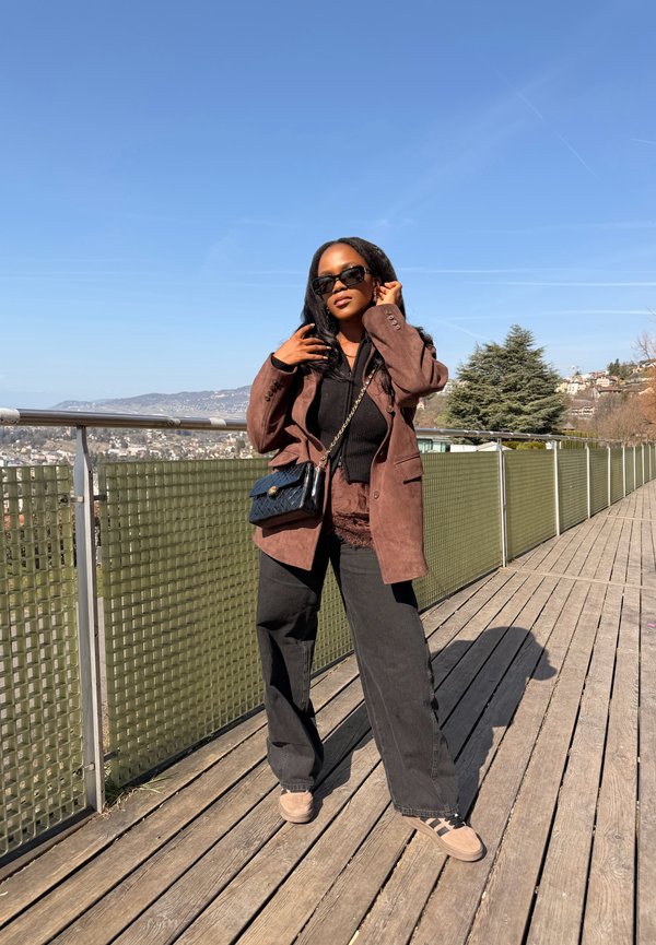 Mujer con gafas de sol, blazer marrón, top negro y pantalones anchos negros, de pie en una terraza de madera con un paisaje urbano y árboles al fondo.