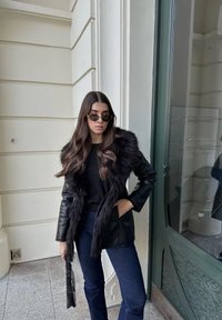 Črna usnjena jakna z umetnim krznom na ovratniku, združena s črno majico in temno moderimi širokimi kavbojkami, dopolnjena z okroglimi sončnimi očali.