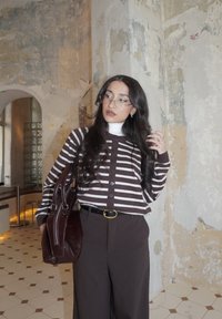 Femme aux longs cheveux foncés portant un cardigan rayé et des lunettes, tenant un sac en cuir foncé, debout dans une pièce ancienne avec un sol carrelé.