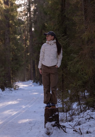 Persona de pie sobre un tocón de árbol en un bosque nevado, vistiendo una chaqueta blanca acolchada, pantalones marrones y un gorro oscuro. Árboles verdes de fondo.