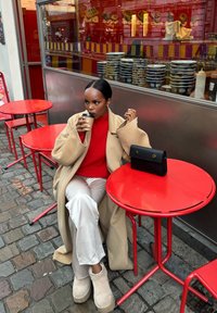 Jasnobeżowy wełniany płaszcz, czerwony sweter, białe spodnie i beżowe buty. Czarna torebka na czerwonym metalowym stole na tle restauracji sushi.