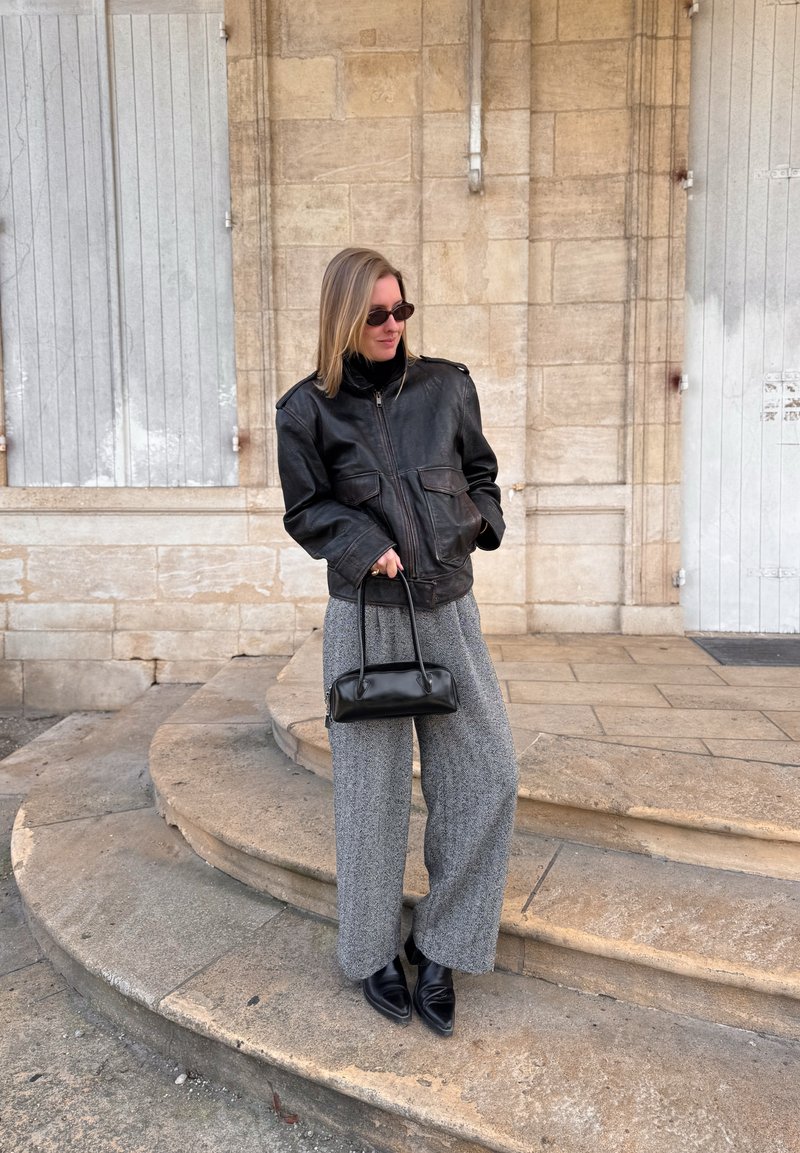 Black leather jacket with zip closure and flap pockets, paired with grey wide-leg trousers and black pointed-toe shoes. Includes a small black handbag.