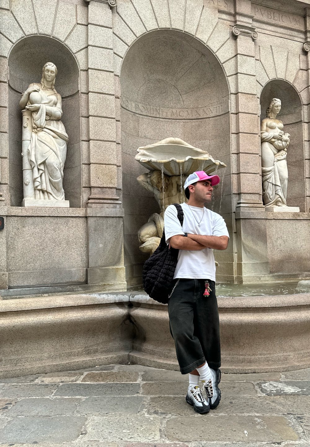 Fuente de rodillas con intrincadas tallas en piedra; figura con camisa blanca, pantalones de pierna ancha negros y un bolso negro texturizado posa cerca.