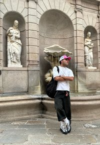 Fuente de rodillas con intrincadas tallas en piedra; figura con camisa blanca, pantalones de pierna ancha negros y un bolso negro texturizado posa cerca.