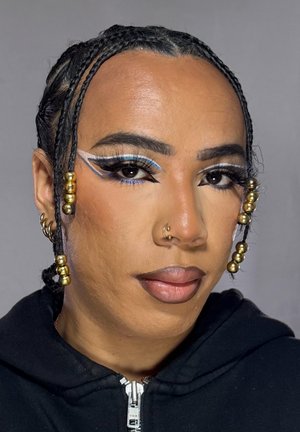 Persona con cabello trenzado adornado con cuentas doradas, intenso delineado de ojos azul y blanco con efecto ala, piercing en la nariz y vistiendo una sudadera negra con cremallera.