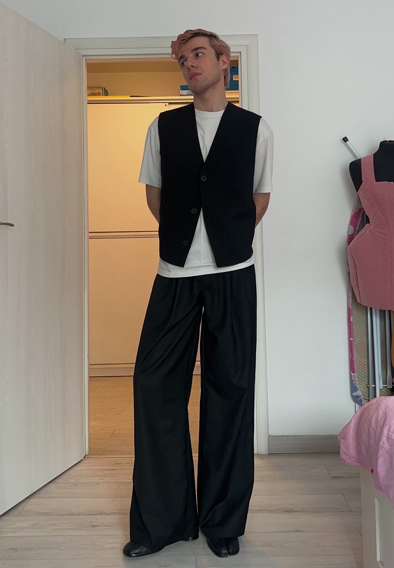 Jeune personne aux cheveux roses portant un t-shirt blanc, un gilet noir boutonné et un pantalon large noir, debout à l'intérieur près d'une porte ouverte et d'un mannequin de couture.