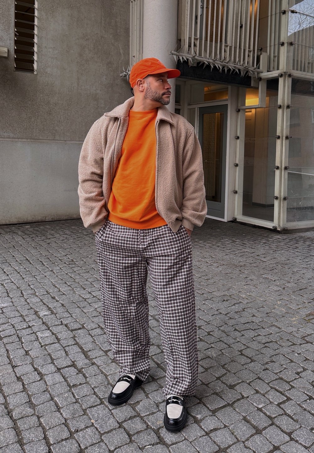 Μπεζ φλις μπουφάν, πορτοκαλί φούτερ, καρό καφέ παντελόνι, μαύρα και λευκά παπούτσια, πορτοκαλί καπέλο. Στέκεται σε επιφάνεια από κυβόλιθους, με σύγχρονη αρχιτεκτονική στο παρασκήνιο.