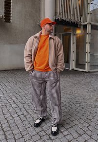 Μπεζ φλις μπουφάν, πορτοκαλί φούτερ, καρό καφέ παντελόνι, μαύρα και λευκά παπούτσια, πορτοκαλί καπέλο. Στέκεται σε επιφάνεια από κυβόλιθους, με σύγχρονη αρχιτεκτονική στο παρασκήνιο.