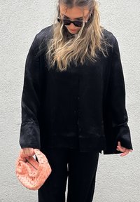 Czarna, oversize koszula z guzikami, z szerokimi rękawami w miękkiej fakturze. Modelka trzyma plecioną torbę w jasnoróżowym kolorze, na neutralnym tle.