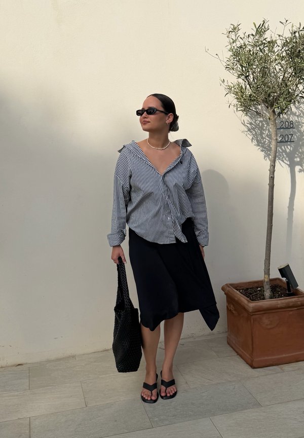 Woman wearing black sunglasses, striped shirt, black skirt, and sandals stands holding a black bag beside a potted tree against a plain wall.