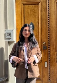 Brun suedjakke over en hvid skjorte, grå bukser og holder solbriller; står nær en trædør med en dørklokke og beslag.