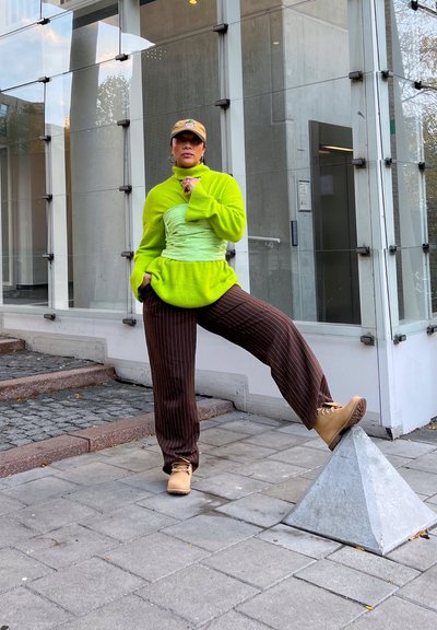 Sudadera verde neón con un diseño asimétrico, superpuesta sobre una tela verde claro, combinada con pantalones marrones de rayas y botas color beige.