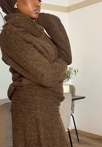 Pull marron tricoté avec un col montant, présentant un tissu texturé et doux. Le modèle pose avec les mains près du visage, mettant en valeur le design du vêtement.