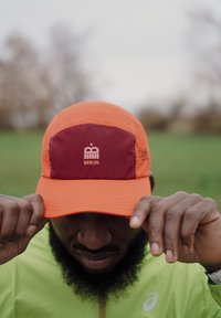 Caps i oransje stoff med en brunrød frontpanel, med en "Berlin"-logo. Designet inkluderer en rundet brem og synlige sømdetaljer.