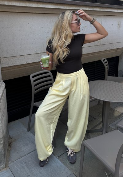 Pantalones amarillos de cintura alta, con pliegues y una textura suave, combinados con una blusa negra ajustada y zapatillas gris, sosteniendo una bebida verde.