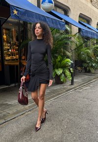 Fekete hosszú ujjú ruha fodrokkal és oldalsó csomós részlettel, bordó lakkbőr magas sarkú cipővel és hozzáillő kézitáskával, utcán sétálva.