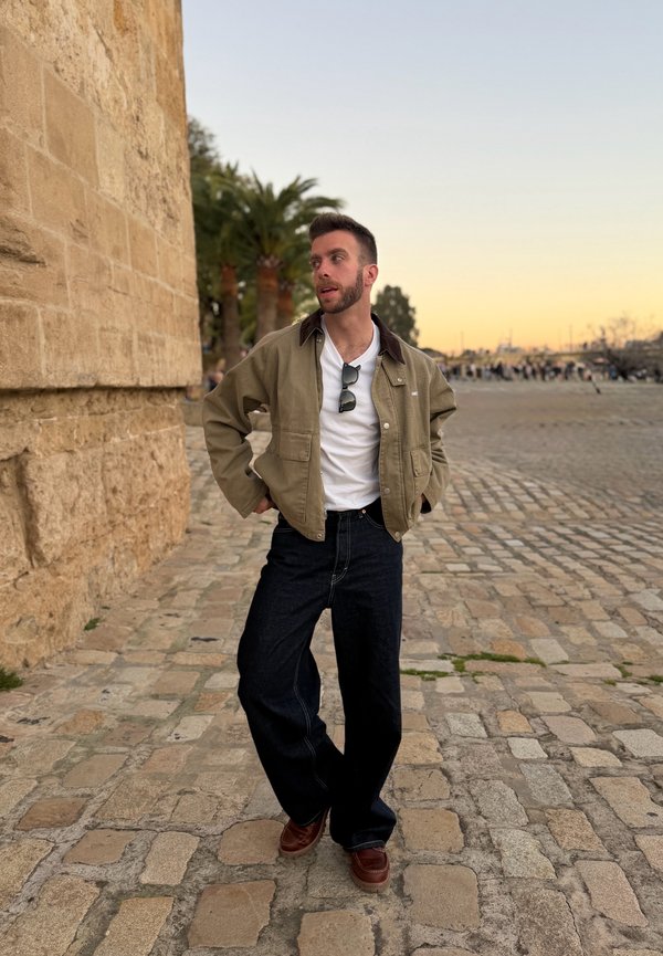 Joven con chaqueta marrón claro y camisa blanca de pie sobre adoquines cerca de un muro de piedra, palmeras y un cielo al atardecer.